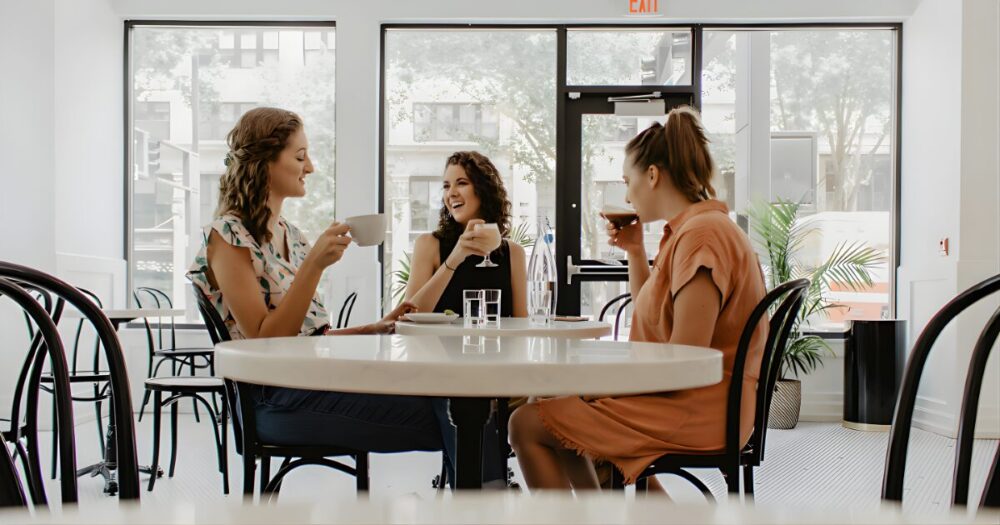 明るいカフェでテーブルを囲み会話を楽しむ三人の女性。女三人の人間関係と力学のバランスを象徴するイメージ