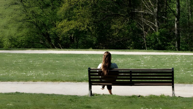 公園のベンチに一人で座り、緑の芝生と木々を前に静かに過ごす女性の後ろ姿
