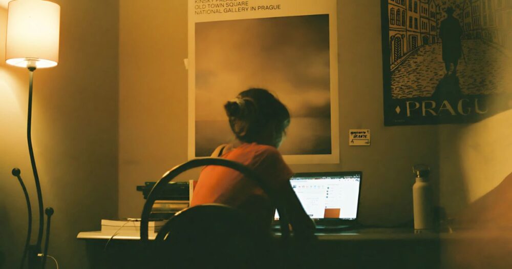 暖かな照明の部屋で、ノートパソコンに向かう女性の後ろ姿。デスクに座り、画面を見つめている。